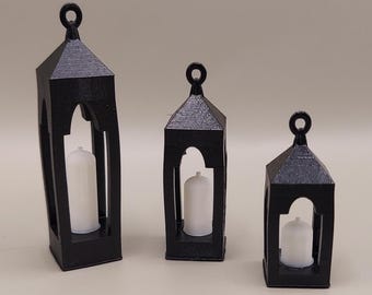 May include: Three black decorative lanterns of different sizes. Each lantern has a rectangular or trapezoidal shape with an arched opening and a small loop at the top. A white candle is inside each lantern.