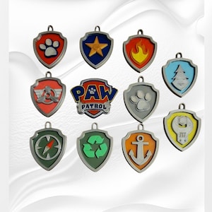 Könnte beinhalten: Ein Set mit 12 Abzeichen der Paw Patrol-Charaktere, jedes mit dem Symbol und der Farbe eines anderen Charakters. Die Abzeichen sind aus Metall und haben eine glänzende Oberfläche. Zu den Charakteren gehören Chase, Marshall, Skye, Rubble, Rocky, Zuma, Everest, Tracker und das Paw Patrol-Logo.