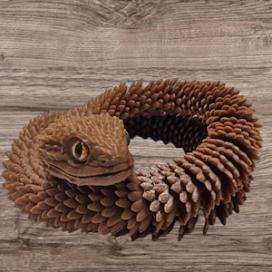 Könnte beinhalten: Ein braunes, schuppiges, schlangenartiges Wesen, das sich zu einem Kreis zusammenrollt. Das Wesen hat einen strukturierten, schuppigen Körper und einen kleinen, spitzen Kopf mit gelben Augen.