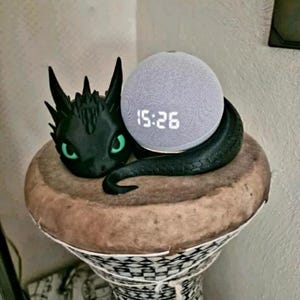 Könnte beinhalten: Eine schwarze Drachenfigur mit grünen Augen ist um einen grauen Smart Speaker mit einer Digitaluhr, die 15:26 anzeigt, gewickelt. Der Lautsprecher steht auf einem braunen Holzsockel mit einem schwarz-weißen geometrischen Muster.