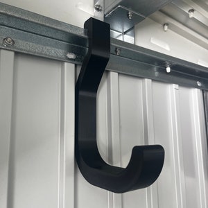 Może przedstawiać: Czarny plastikowy hak w kształcie litery J zamontowany na metalowej belce. Hak jest przeznaczony do zawieszania przedmiotów na suficie.