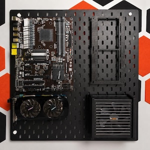 May include: A black pegboard wall mount displaying a computer motherboard, a dual-fan graphics card, a power supply unit with a visible fan, and a rectangular frame. The components are arranged on the perforated surface for hardware organization.