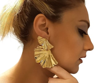 May include: Gold-colored statement earrings with a layered, fan-like design. The earrings have a textured, reflective surface and are worn by a person with blonde hair. The earrings are a bold accessory.
