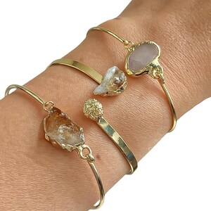 May include: Four gold bangles with different gemstone charms. The charms include a clear quartz, a pink quartz, a white shell, and a brown crystal.