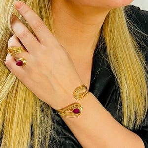 Könnte beinhalten: Ein goldfarbenes Armband und ein Ringset mit einem burgunderfarbenen Edelstein. Das Armband ist offen und hat ein geschwungenes Design. Der Ring ist ein dünner Band mit einem einzigen Edelstein.