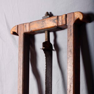 Pode incluir: Uma moldura de madeira antiga com uma lâmina de serra de metal suspensa no centro. A moldura é feita de madeira castanha clara envelhecida, e a lâmina de serra mostra sinais de ferrugem. O design sugere uma ferramenta para marcenaria ou artesanato similar.
