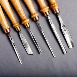 Puede incluir: Un conjunto de cinceles para tallar madera con mangos de madera clara y hojas de metal. Los cinceles están dispuestos en fila, mostrando sus diferentes formas y tamaños de hojas. Los mangos tienen virolas de latón.