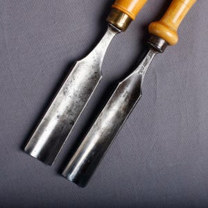 May include: Two antique wood chisels with metal blades and wooden handles. The blades are silver and show signs of wear. The handles are light brown with brass ferrules. The tools are likely used for woodworking or carving.
