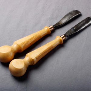 K&ouml;nnte beinhalten: Zwei Holzschnitzgouge mit hellbraunen Griffen und dunklen Metallklingen. Die Griffe haben ein abgerundetes Ende und einen geriffelten Abschnitt f&uuml;r den Griff. Die Klingen sind gebogen. Die Werkzeuge sind vor einem grauen Hintergrund platziert.