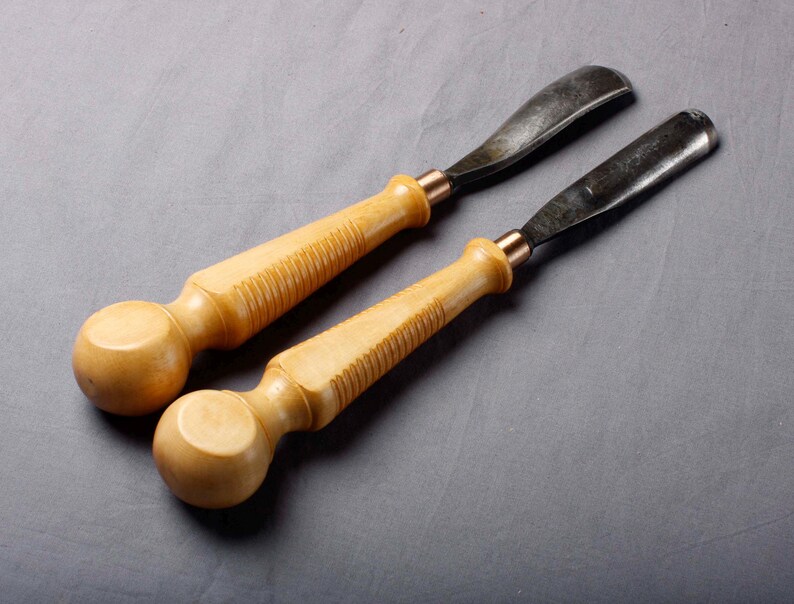 K&ouml;nnte beinhalten: Zwei Holzschnitzgouge mit hellbraunen Griffen und dunklen Metallklingen. Die Griffe haben ein abgerundetes Ende und einen geriffelten Abschnitt f&uuml;r den Griff. Die Klingen sind gebogen. Die Werkzeuge sind vor einem grauen Hintergrund platziert.