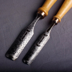 Peut inclure: Deux ciseaux à bois avec des manches brun clair et des lames en métal. Les lames ont un aspect sombre et texturé et sont inclinées au niveau du tranchant. Les ciseaux sont posés sur un fond gris.