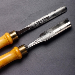 以下が含まれることがあります： 金属刃と明るい黄色の柄が付いた、2つのヴィンテージ木彫りノミ。刃には「PEUGEOT FRERES」と「A. ZONCA FOND.」の文字が刻まれています。ツールは灰色の背景に配置されています。