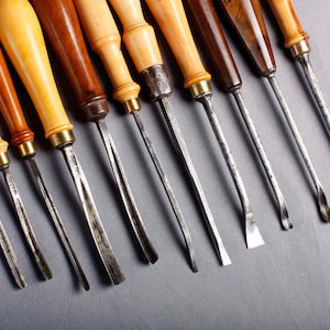 Può includere: Una collezione di utensili per intaglio del legno con manici in legno e lame in metallo. I manici variano di colore dal marrone chiaro allo scuro e le lame hanno forme diverse, tra cui dritte, curve e angolate.