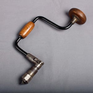 Puede incluir: Un taladro manual vintage con mango de madera y marco de metal negro. El taladro tiene un portabrocas para sujetar brocas y un pomo de madera para girar. Esta herramienta de perforación manual se utiliza para trabajos de carpintería u otros proyectos.