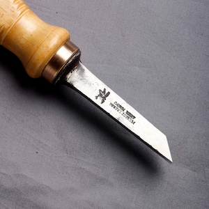 May include: A wood carving knife with a light wooden handle and a metal blade. The blade has text and a small emblem. The handle has carved details and a metal ferrule. The tool is designed for woodworking and is approximately 15 cm long.