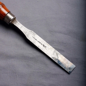 May include: A close-up shot of a vintage chisel. It has a wooden handle and a metal blade. The blade is marked with "PEUGEOT FRERES +" and a logo. The handle is dark brown, and the blade is silver.