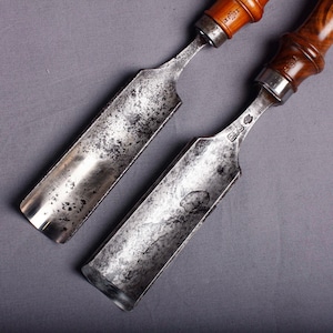 Puede incluir: Dos gubias metálicas antiguas con mangos de madera. Las hojas muestran signos de edad y uso, con una forma curvada y un acabado plateado. Los mangos son de color marrón oscuro.