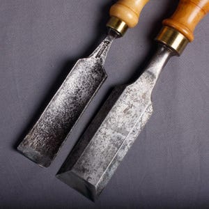 Op de afbeelding: Twee antieke houtbeitels met metalen bladen. De beitels hebben lichtbruine houten handvatten met messing hulzen. De bladen zijn dof zilverkleurig en vertonen tekenen van slijtage. De gereedschappen zijn naar de kijker gericht.