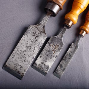 May include: Three antique wood chisels with steel blades and wooden handles. The chisels are arranged diagonally, showcasing their varying sizes and the "WARD CAST STEEL" markings. The handles are a light brown color, and the blades show signs of wear.