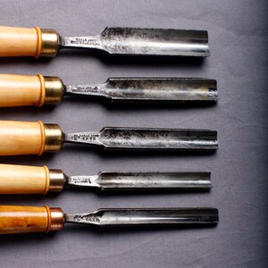 Puede incluir: Un juego de cinco cinceles para tallar madera con mangos de madera y hojas de metal. Las hojas son de metal oscuro y envejecido, y los mangos varían en color, desde madera clara hasta oscura. Los cinceles están dispuestos en fila, mostrando sus diferentes formas y tamaños.