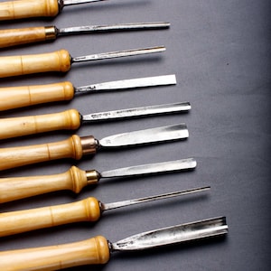 May include: A selection of wood carving tools, featuring wooden handles and metal blades. The tools are displayed diagonally against a dark grey backdrop. The blades are of different shapes, including pointed, flat, and curved.