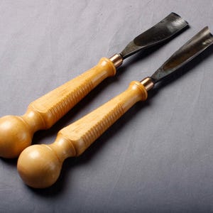 K&ouml;nnte beinhalten: Zwei Holzschnitzgouge mit hellbraunen Holzgriffen und gebogenen, dunklen Metallklingen. Die Griffe haben ein knollenf&ouml;rmiges Ende und einen geriffelten Griffbereich. Die Metallklingen sind mit kupferfarbenen Zwingen befestigt.