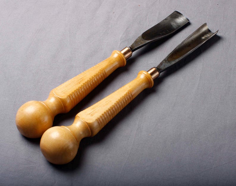 K&ouml;nnte beinhalten: Zwei Holzschnitzgouge mit hellbraunen Holzgriffen und gebogenen, dunklen Metallklingen. Die Griffe haben ein knollenf&ouml;rmiges Ende und einen geriffelten Griffbereich. Die Metallklingen sind mit kupferfarbenen Zwingen befestigt.