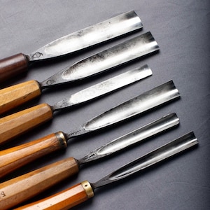 Może przedstawiać: Kolekcja dłut do obróbki drewna z drewnianymi uchwytami i metalowymi ostrzami. Dłuta ułożone są po przekątnej na ciemnym tle. Uchwyty są w kolorze jasnobrązowym, a ostrza srebrne.