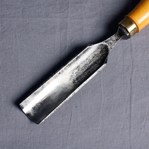May include: A close-up of a metal wood chisel with a wooden handle. The chisel is silver with a slightly dull finish. The handle is a light brown wood with a brass ferrule.