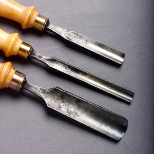 Peut inclure: Trois ciseaux à bois avec des manches en bois clair et des lames en métal. Les lames sont en métal sombre vieilli, et les manches ont une virole en laiton. Les outils sont disposés en diagonale sur un fond gris.