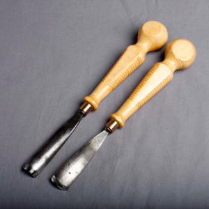 K&ouml;nnte beinhalten: Zwei Holzschnitzbeitel mit hellen Holzgriffen und Metallklingen. Die Griffe haben ein rundes Ende und einen geriffelten Griff. Die Klingen sind dunkelmetallfarben. Auf grauem Hintergrund.