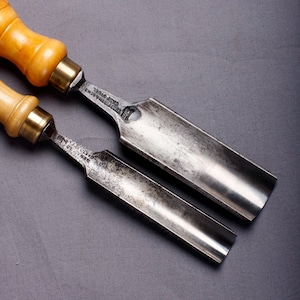 May include: Two antique wood carving gouges with wooden handles and metal blades. The handles are light brown with brass ferrules. The blades are curved and show signs of wear, indicating their age and use. The tools are likely used for woodworking.