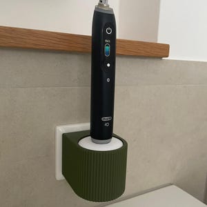 Könnte beinhalten: Eine schwarze elektrische Zahnbürste Oral-B iO befindet sich in einem grünen Halter an einer Badezimmerwand. Die Zahnbürste zeigt einen Batterieladestand von 94 %. Der Halter ist olivgrün mit geripptem Design.