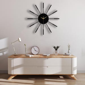 Puede incluir: Un reloj negro y dorado con un dise&ntilde;o moderno. El reloj tiene una esfera negra con agujas doradas y un marco negro con detalles dorados. El reloj est&aacute; colgado en una pared blanca sobre una c&oacute;moda blanca con dos cajones.