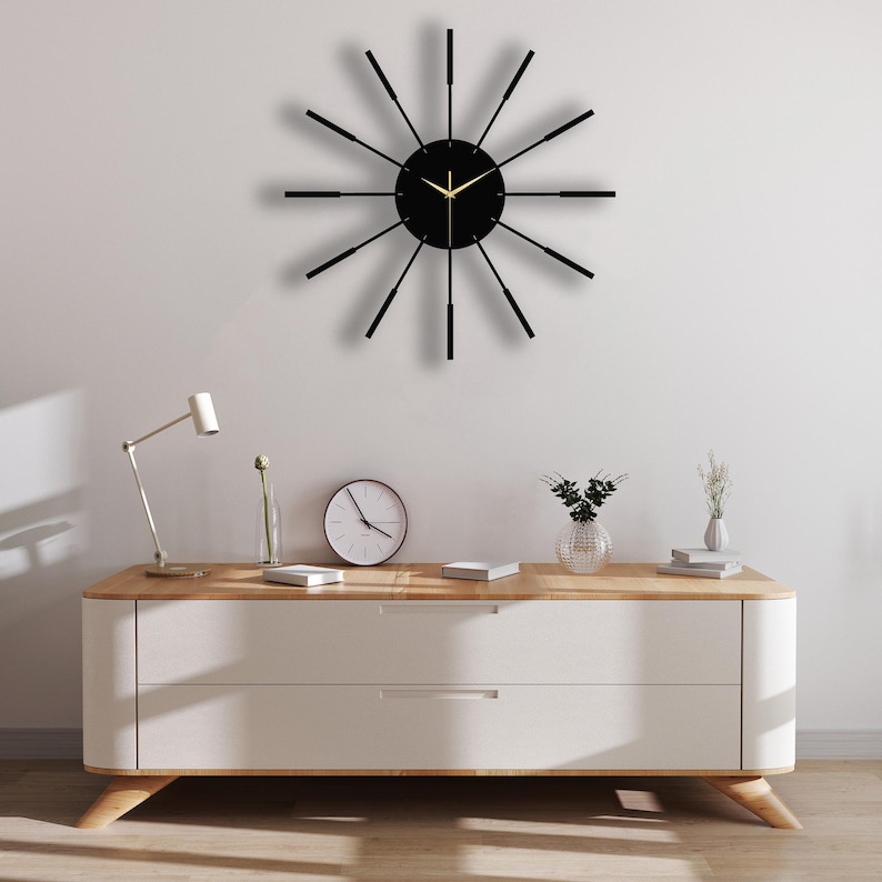 Puede incluir: Un reloj negro y dorado con un dise&ntilde;o moderno. El reloj tiene una esfera negra con agujas doradas y un marco negro con detalles dorados. El reloj est&aacute; colgado en una pared blanca sobre una c&oacute;moda blanca con dos cajones.