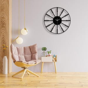 May include: A black metal wall clock with a large, open face and thin, black hands. The clock is hanging on a white wall in a living room setting with a pink armchair and a side table.