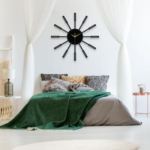 May include: A black and gold clock with a unique design featuring long, thin spokes radiating from the centre. The clock is mounted on a white wall above a bed with a green blanket and patterned pillows.