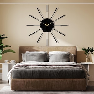 Puede incluir: Un reloj de pared moderno negro y dorado con largas manecillas negras delgadas y una esfera negra. El reloj est&aacute; colgado en una pared sobre una cabecera marr&oacute;n con ropa de cama gris.