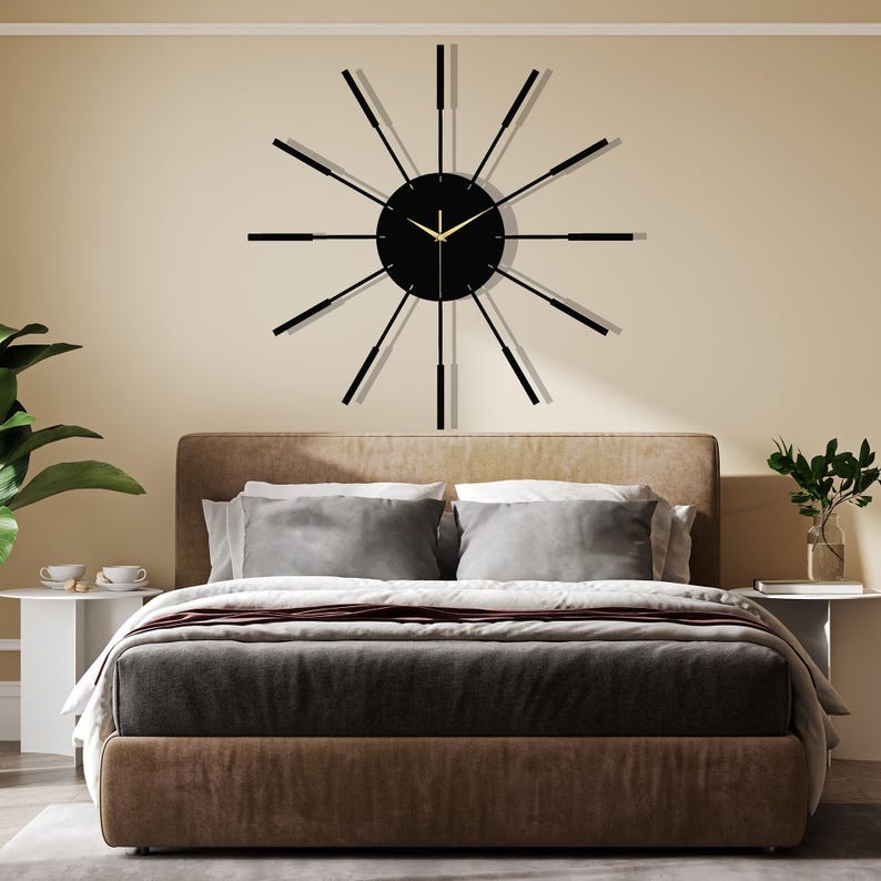 Puede incluir: Un reloj de pared moderno negro y dorado con largas manecillas negras delgadas y una esfera negra. El reloj est&aacute; colgado en una pared sobre una cabecera marr&oacute;n con ropa de cama gris.