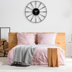 Puede incluir: Un reloj de metal negro con una gran esfera circular y radios finos y radiantes. El reloj está colgado en una pared blanca sobre una cama rosa con una cabecera de madera y una manta gris.