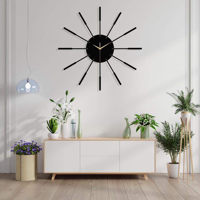 Puede incluir: Un reloj negro y dorado con largas agujas negras finas y una esfera negra. El reloj est&aacute; colgado en una pared blanca sobre un armario blanco con dos cajones. Hay plantas en macetas a ambos lados del armario.