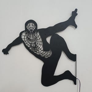 Può includere: Arte da parete in metallo nero che rappresenta un supereroe con un costume a rete, in posa in una posa d'azione dinamica. L'opera d'arte è illuminata da dietro, creando un effetto di silhouette drammatico.