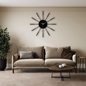 Puede incluir: Un reloj negro y dorado con un dise&ntilde;o de rayos de sol cuelga de una pared beige sobre un sof&aacute; beige con almohadas marrones. Una mesa de caf&eacute; marr&oacute;n se encuentra frente al sof&aacute;.