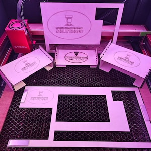 May include: A laser cutter with a pink honeycomb cutting mat. There are several pieces of wood with the text "LOUD CREATIVE CRAFT SOLUTIONS" on them. The pieces are arranged in a pattern on the mat.