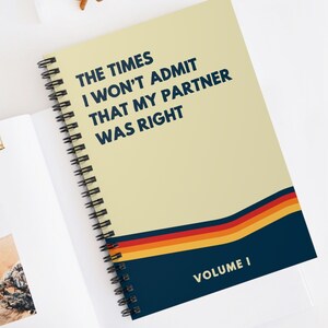 May include: A spiral bound notebook with a yellow cover and blue and red stripes. The text on the cover reads "The Times I Won't Admit That My Partner Was Right" and "Volume I".