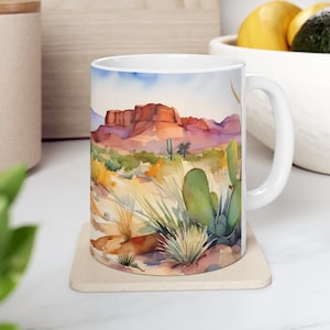 May include: White ceramic mug with a watercolor design of a desert landscape. The design features a red rock formation in the background, with cacti and other desert plants in the foreground.