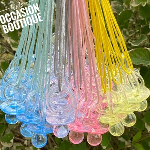 May include: A collection of colorful pacifiers with matching cords. The pacifiers are in shades of blue, pink, yellow, and gray. The text "Kids Occasion Boutique" is in the upper left corner.