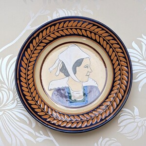 May include: A round ceramic plate featuring a central illustration of a person in profile wearing a white bonnet and a blue and purple garment. The rim is decorated with a repeating brown leaf pattern on a tan background, framed by dark blue borders.