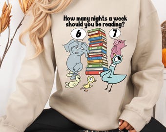 Puede incluir: Sudadera beige con un gráfico colorido. El gráfico presenta animales de dibujos animados, una pila de libros y el texto: "¿Cuántas noches a la semana deberías leer?" con los números 6 y 7.