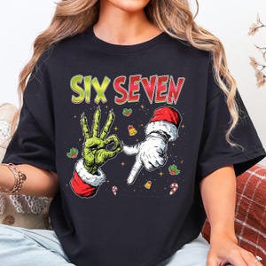 Puede incluir: Camiseta negra con un gr&aacute;fico de dos manos, una verde y otra blanca, formando los n&uacute;meros seis y siete. El texto "SIX SEVEN" est&aacute; en verde y rojo. Los gr&aacute;ficos festivos de acebo, campanas y bastones de caramelo completan el dise&ntilde;o.